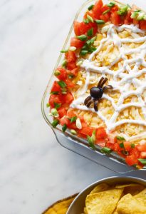 Halloween 7-Layer Lamb Dip topped with shredded cheese, tomatoes, green onions, and a sour cream spider web.