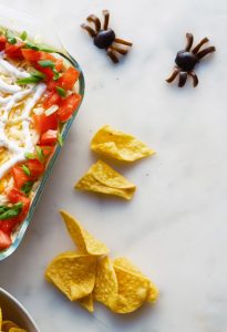 Halloween 7-Layer Dip with a sour cream spider-web design served with tortilla chips.
