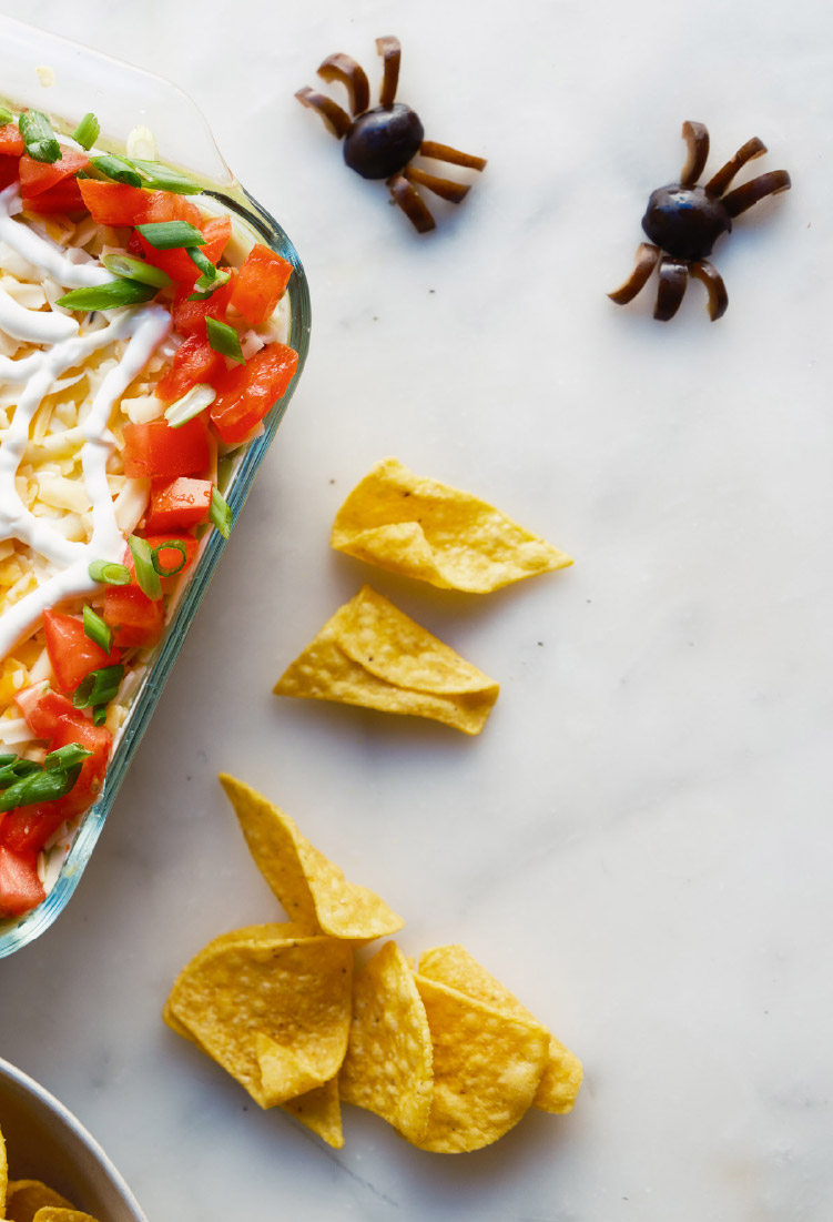 Halloween 7-Layer Dip with a sour cream spider-web design served with tortilla chips.
