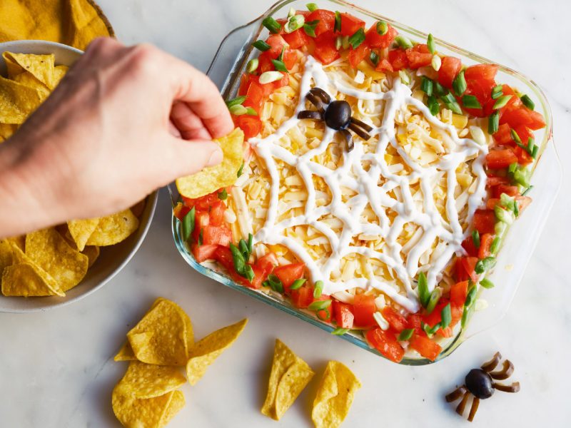 Halloween 7-Layer Dip with spiced American Lamb, cheese, tomatoes, green onions, and a sour cream spider-web design