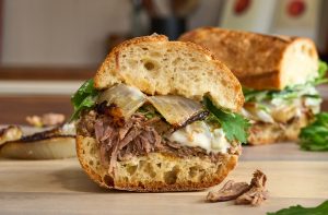 Slow-Cooked American Lamb Sandwich with shredded lamb, charred onions, greens, and creamy jalapeño aioli on a toasted baguette.